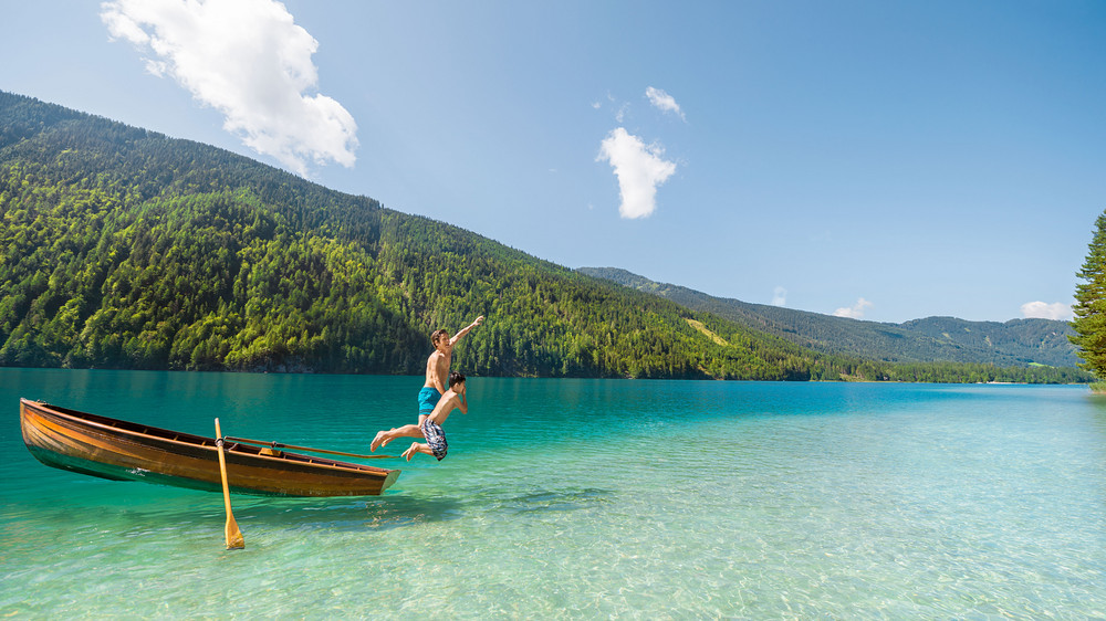 Mann und Junge springen von einem Ruderboot in einen See im Sommerurlaub in Kärnten