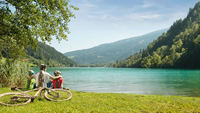 Mann und Kinder sitzen an einem See nach einer Radtour im Radurlaub in Kärnten