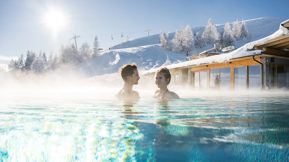 Ein Pärchen genießt die Wärme des Unendlichpools im Winter, im Hintergrund das Skigebiet Gerlitzen Alpe