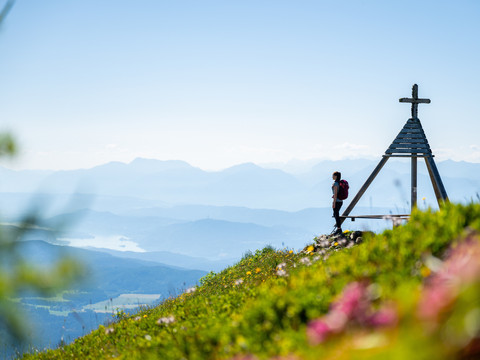 Eine Person steht neben dem Wetterkreuz und blickt in die Ferne. Es ist ein sonniger Sommertag.