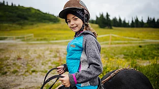 Ein Kind mit Reithelm auf einem Pferd, im Hintergrund blühende Almwiese.